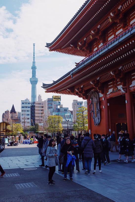 Tourists and students in front of the Senso-ji temple, with the Tokyo Skytree tower visible in the back.