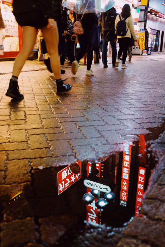 Shop signs reflecting in a small pool of water while people are walking by in Shibuya in Tokyo, Japan.